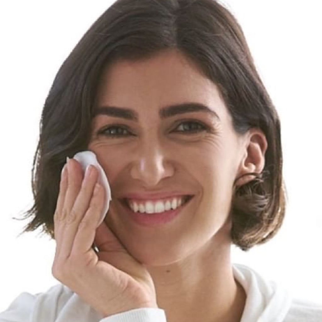 Woman cleaning her face with a white cloth against a plain background